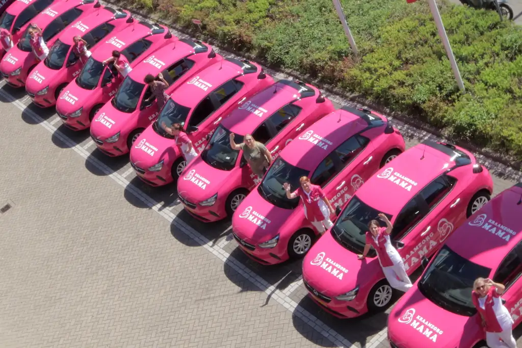Groep enthousiaste kraamverzorgsters van Kraamzorg Mama die vriendelijk zwaaien vanuit hun opvallende roze bedrijfswagen.
